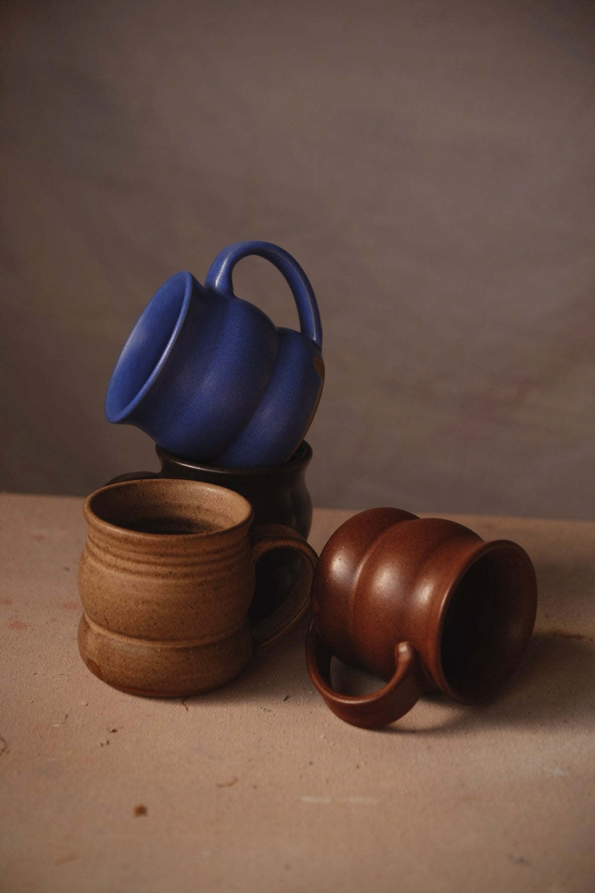 The Wave Mug: four ribbed stoneware mugs stacked and tumbled on a linen-covered table, in cobalt blue, black, sand and rust-brown glazes. Hand thrown by Settle Ceramics in Austin, Texas.